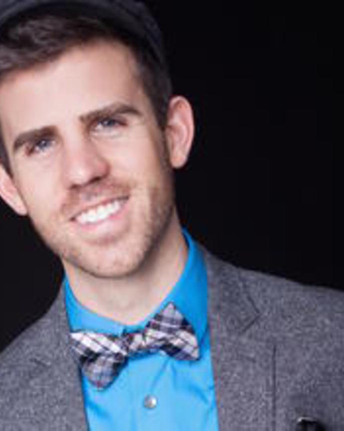 Smiling man in a blue shirt and grey wool jacket, with a plaid bow tie